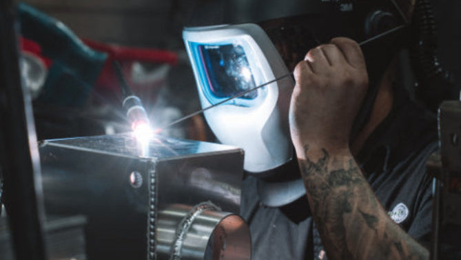 Person welding metal with a welding helmet on, wearing a protective suit.